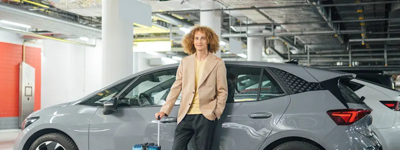junger Mann lehnt mit überkreuzten Beinen und Koffer lässig an einem parkenden Auto in der Tiefgarage
