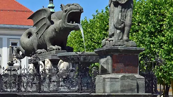 Parken in Klagenfurt: Im Vordergrund ist der Drachenbrunnen zu sehen im Hintergrund befindet sich das Rathaus der Stadt Klagenfurt.