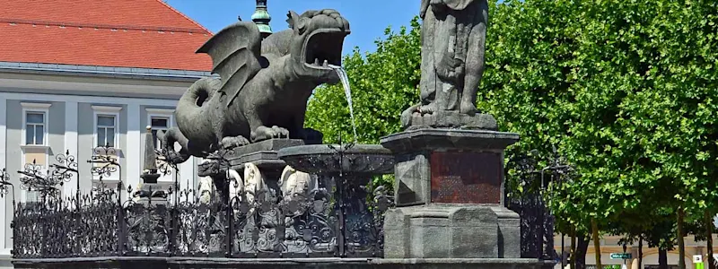Parken in Klagenfurt: Im Vordergrund ist der Drachenbrunnen zu sehen im Hintergrund befindet sich das Rathaus der Stadt Klagenfurt.