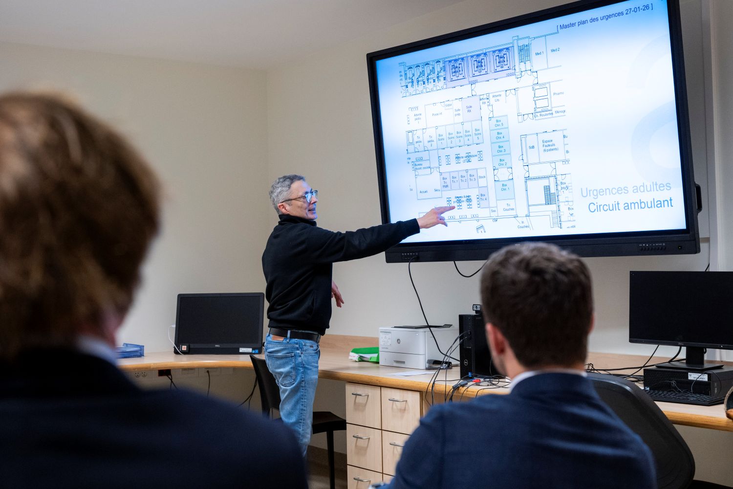 Un représentant de l'hôpital présente le master plan de rénovation du service des urgences du CHU Saint-Pierre à un public, en pointant un plan architectural détaillé affiché sur grand écran.