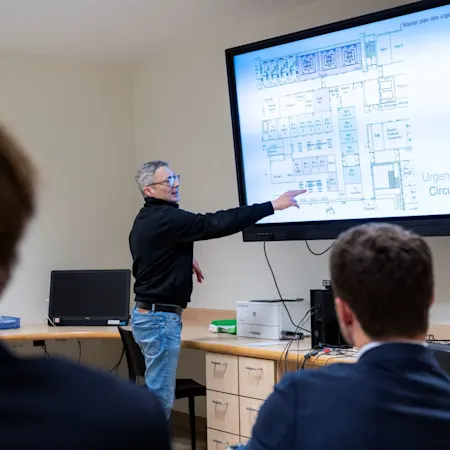 Un représentant de l'hôpital présente le master plan de rénovation du service des urgences du CHU Saint-Pierre à un public, en pointant un plan architectural détaillé affiché sur grand écran.