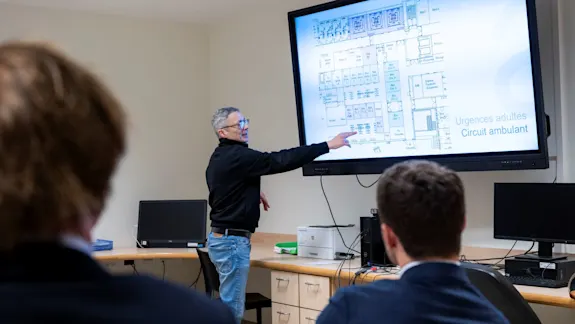 Un représentant de l'hôpital présente le master plan de rénovation du service des urgences du CHU Saint-Pierre à un public, en pointant un plan architectural détaillé affiché sur grand écran.