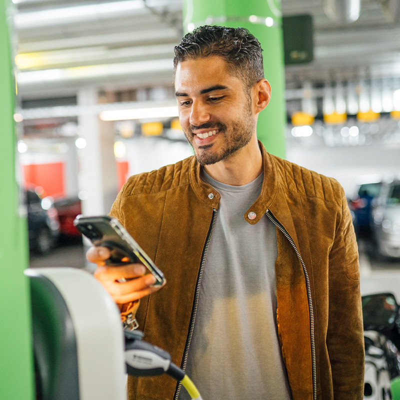 junger Mann steht in der Tiefgarage im Bereich des E-Ladens mit dem Smartphone vor der Wallbox
