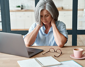 Eine grauhaarige besorgte Frau sitzt am Laptop und fasst sich ihren Nacken mit beiden Händen