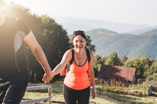 Lächelnde Frau in Sportkleidung geht einen Bergpfad entlang. Sie hält einen Mann an der Hand. Im Hintergrund sind Berge.  