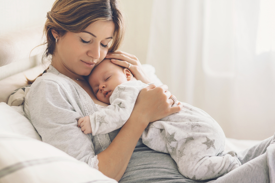 Eine Frau in halbliegender Position mit geschlossenen Augen. Sie umarmt einen schlafenden Säugling in grauer Kleidung.
