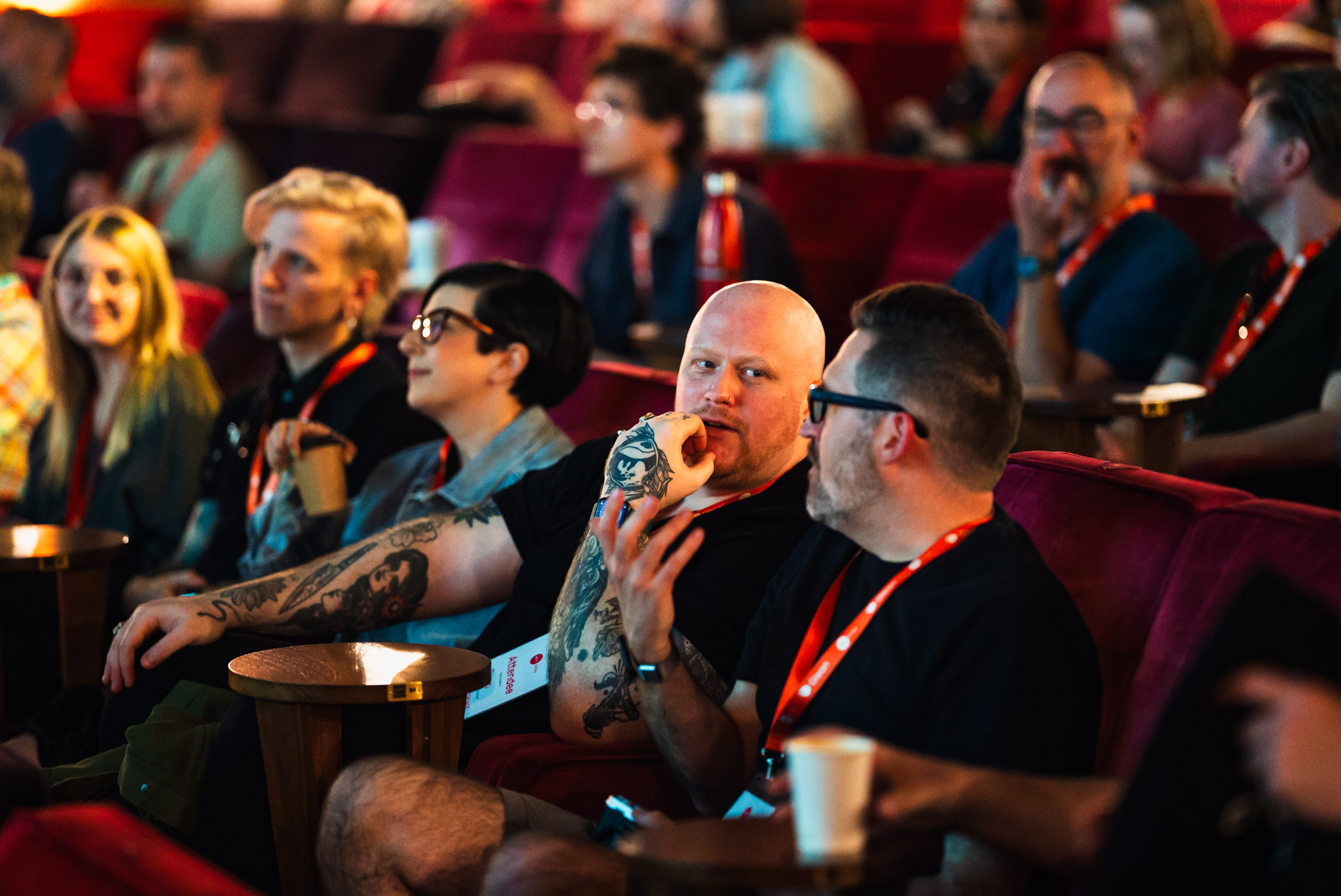 A row of conference attendees are photographed whilst watching a speaker on the stage. Some are looking at the camera, some at the stage intently, and others are talking about what they are seeing in hushed voices.