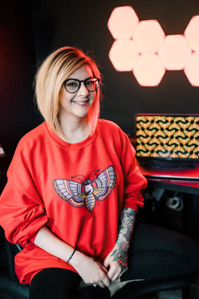 Salma is sitting cross-legged on a stool, wearing a red panthermoth sweater. Her laptop is to the right, below a block of nano leaf lights on a black wall.