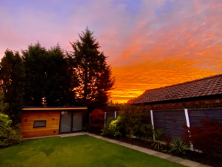 My wooden chalet style office at the bottom of the garden, with a very orange sunrise backdrop fading into a purple hue.