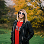 A headshot of Salma in a field with autumnal trees behind her. She is wearing a leather jacket, red moth sweatshirt, and sunglasses.