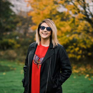 A headshot of Salma in a field with autumnal trees behind her. She is wearing a leather jacket, red moth sweatshirt, and sunglasses.