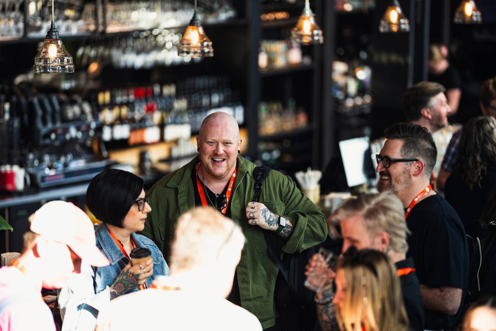A group of people are photographed from an elevated angle against a blurred backdrop of a bar. The group are holding coffees, back packs, and laughing. The sun beats down from the glass in the ceiling.