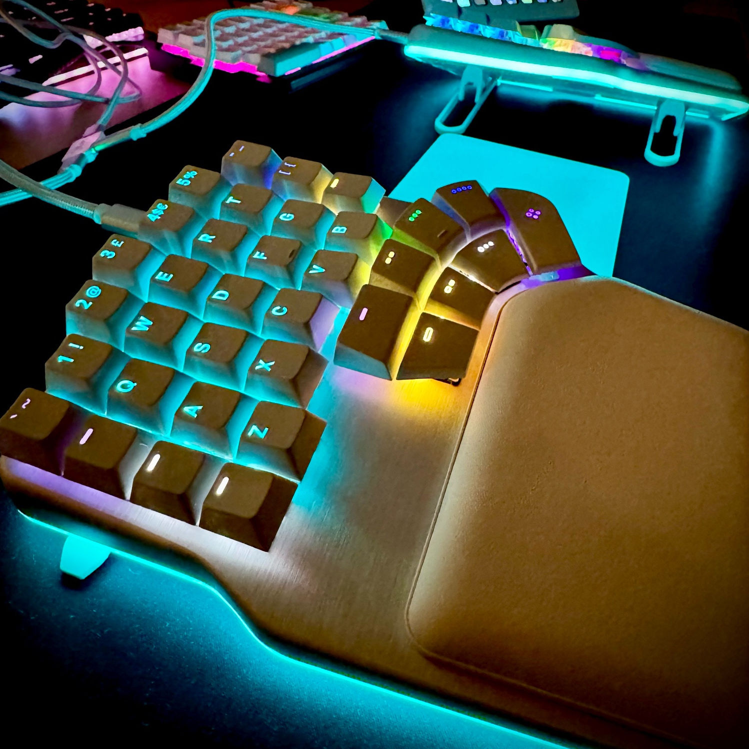 The left half of a white and silver dygma defy keyboard, in the dark on a black desk matt, lit up with rainbow RGB. There’s another split keyboard behind it, lit purple and a white stream deck in the background.