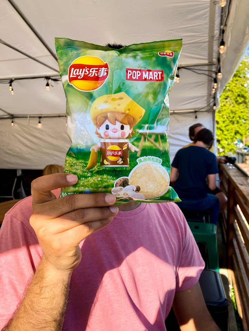 My fiancé holding a bag of chips in front of his face at a beer garden. On the bag is an illustration of a little boy wearing a mushroom cap hat.