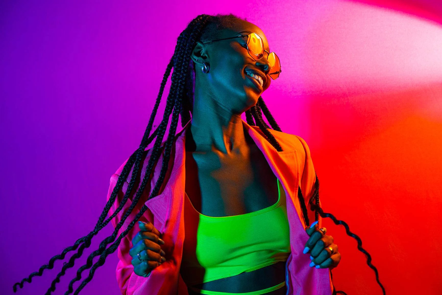 A woman with long braided hair is wearing a neon-colored top and an open jacket against a backdrop of pink and red RGB lighting.