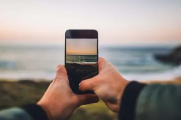 Hands holding a smartphone showing a photo of the sea with a sunset, blurred beach in the background.