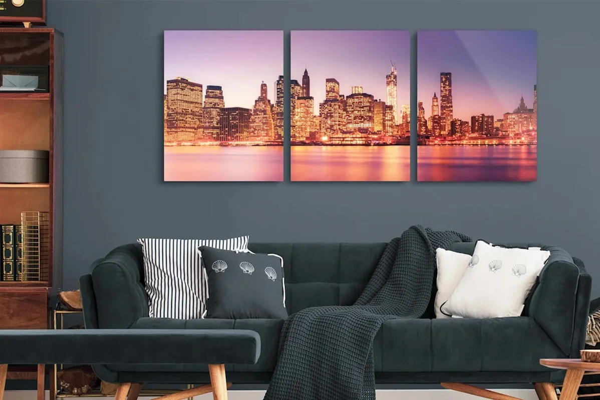 Trittico con silhouette urbana illuminata al crepuscolo sopra un divano con cuscini e coperta in un salotto moderno.