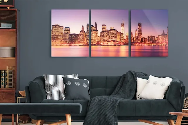 Trittico con silhouette urbana illuminata al crepuscolo sopra un divano con cuscini e coperta in un salotto moderno.