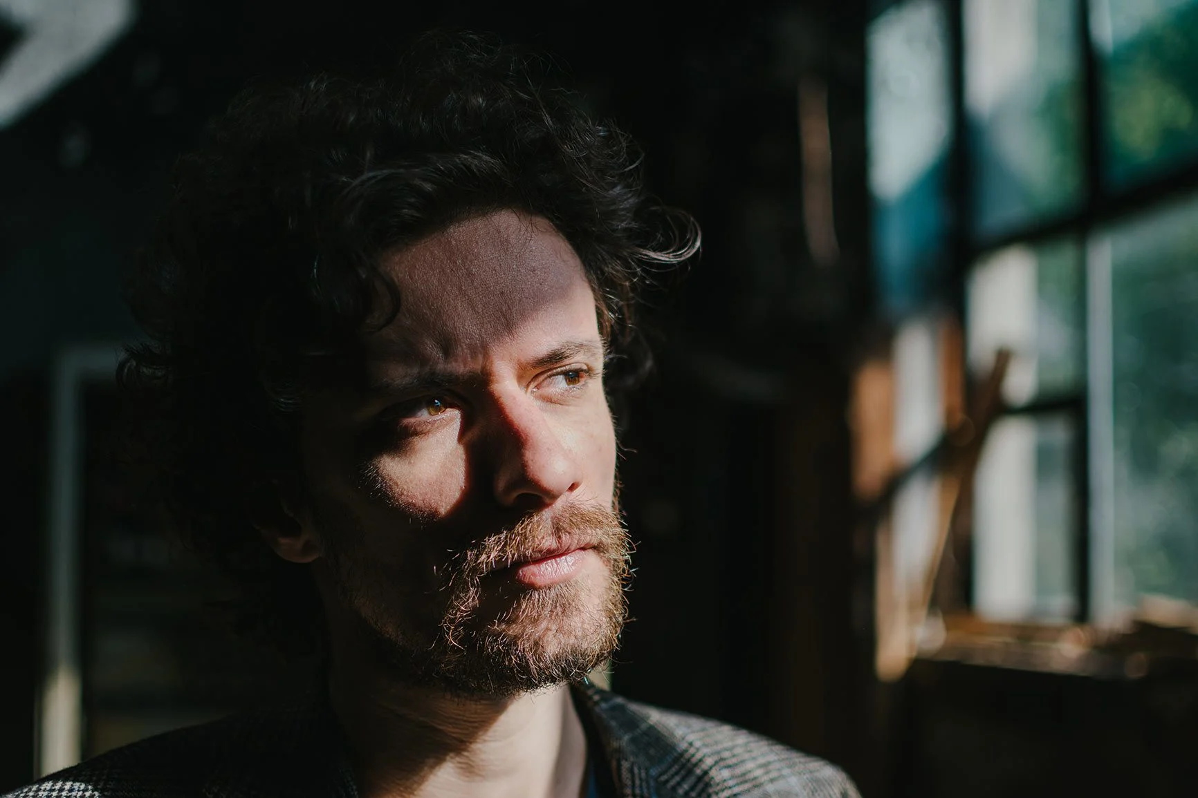 Portrait eines Mannes mit lockigem Haar und Bart vor einem Fenster mit Gitterstruktur in dunklem Raum.