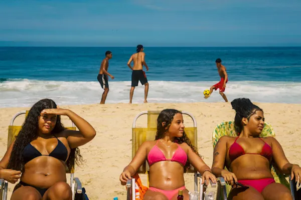 A sunny beach scene by the ocean. In the foreground, three young women in bikinis sit in beach chairs, looking in different directions. In the background, near the shoreline of the receding waves, three young men play football on the light sand under a clear blue sky.