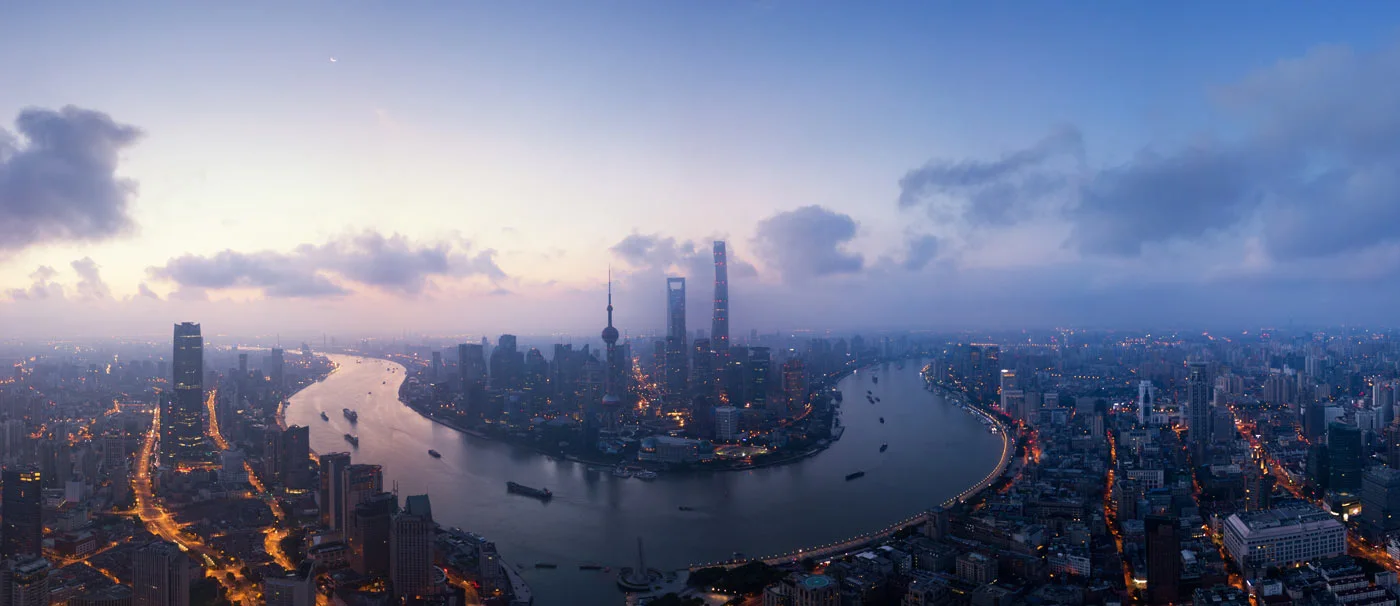Panorama of Shanghai's skyline at dusk with the Huangpu River, clouds, and illuminated skyscrapers.