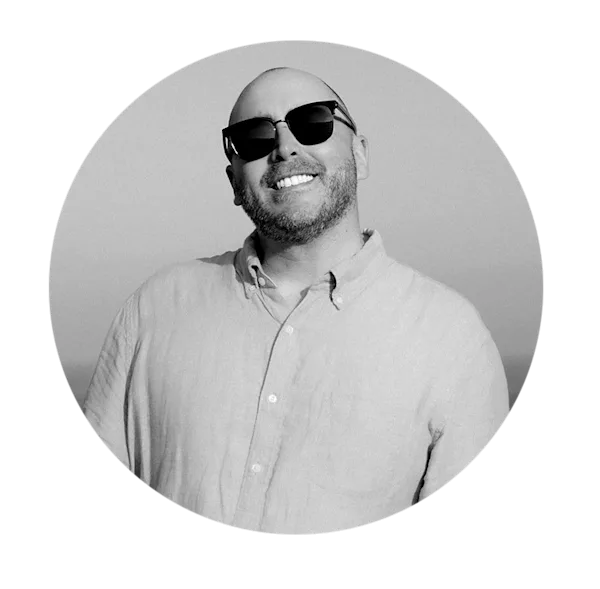 Ein kreisrundes, stimmungsvolles Porträtfoto von James Katsipis vor einem hellen, leicht körnigen Hintergrund. Er ist glatzköpfig, trägt eine Sonnenbrille und ein helles Leinenhemd. Ein breites, authentisches Lächeln prägt das Bild. Das Motiv zeigt ihn im Halbprofil, den Kopf leicht nach hinten geneigt.