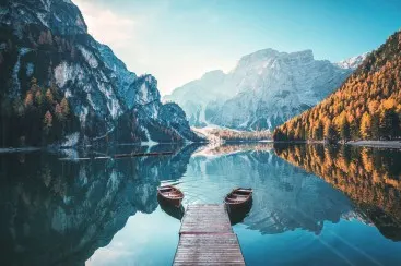 Wooden jetty with two rowboats on a tranquil mountain lake, surrounded by wooded and rocky mountains in autumn.