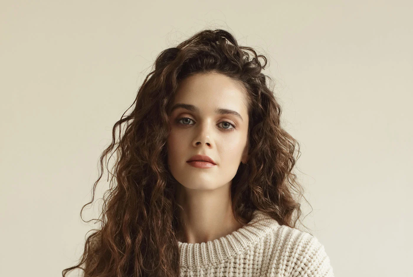 Woman with long, curly brown hair wearing a cream-colored knit sweater.