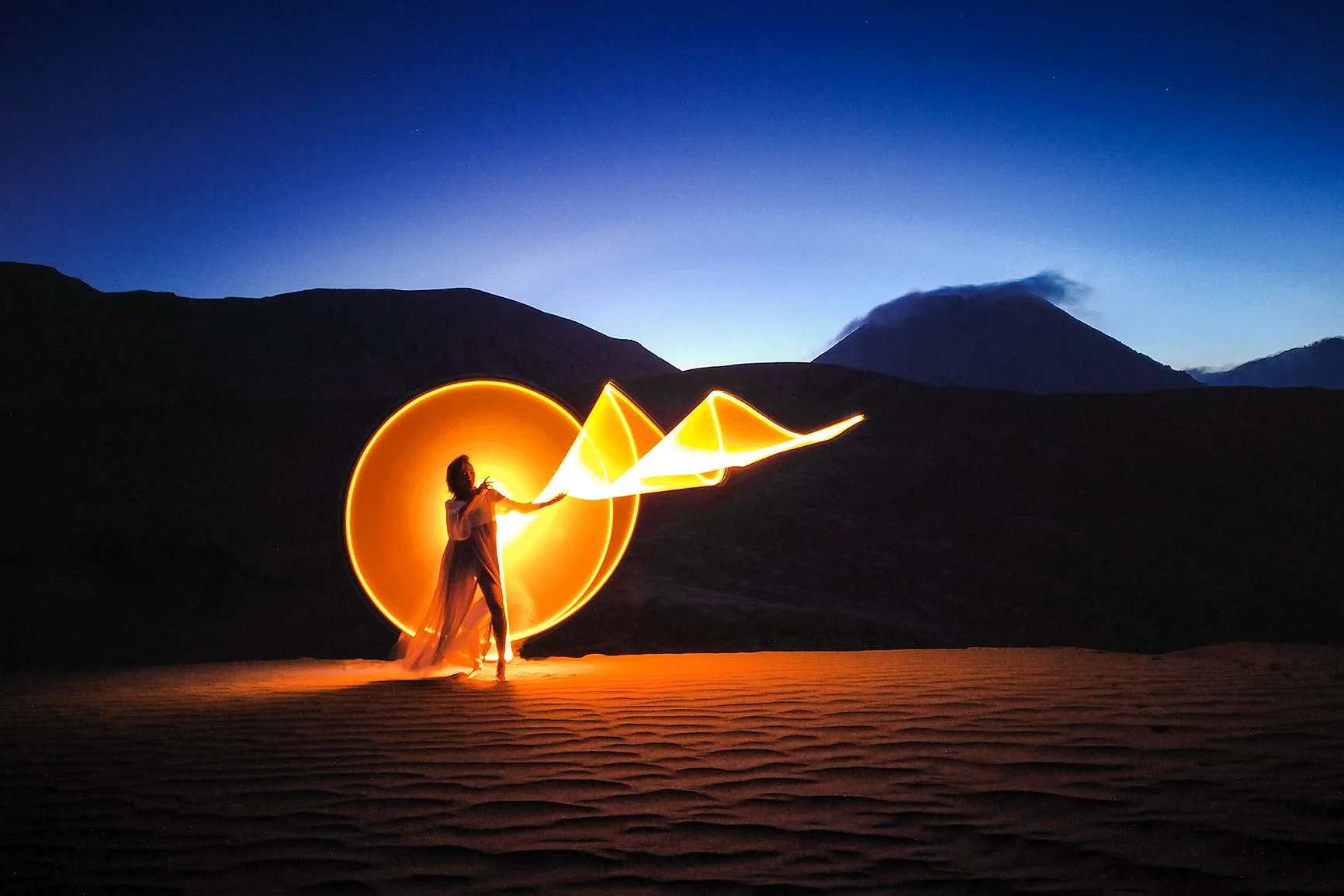 In the desert, a woman uses a light source to create glowing orange circles and wave patterns during a long exposure.
