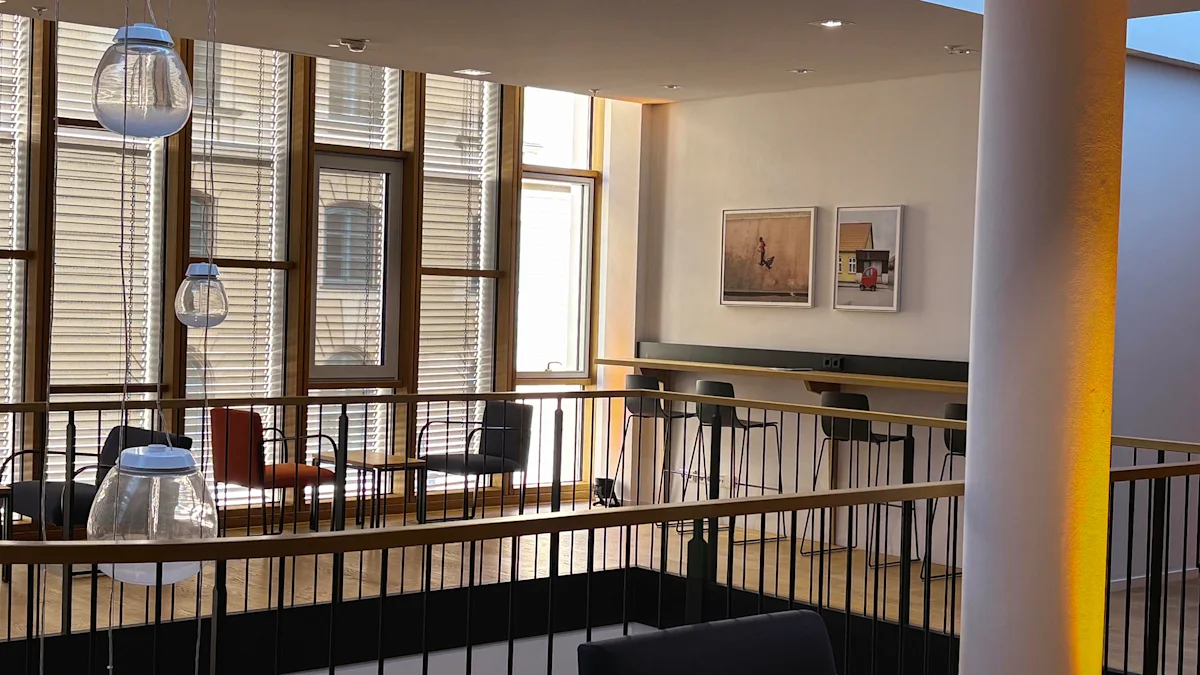 Modern office lounge with glass pendant lights, bar seating, large windows with blinds, and two framed artworks on a white wall.