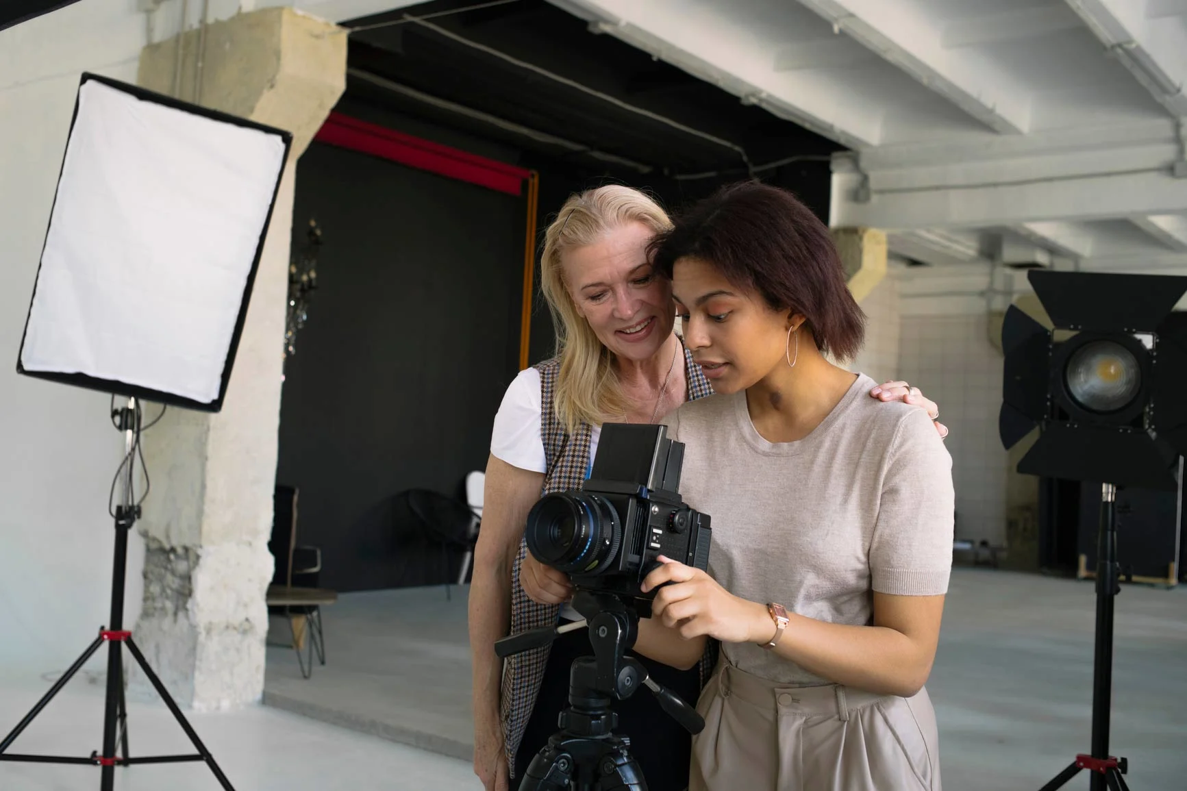 Two women stand in a photo studio and look together at the camera settings on a large camera. Studio lights and equipment are visible in the background.