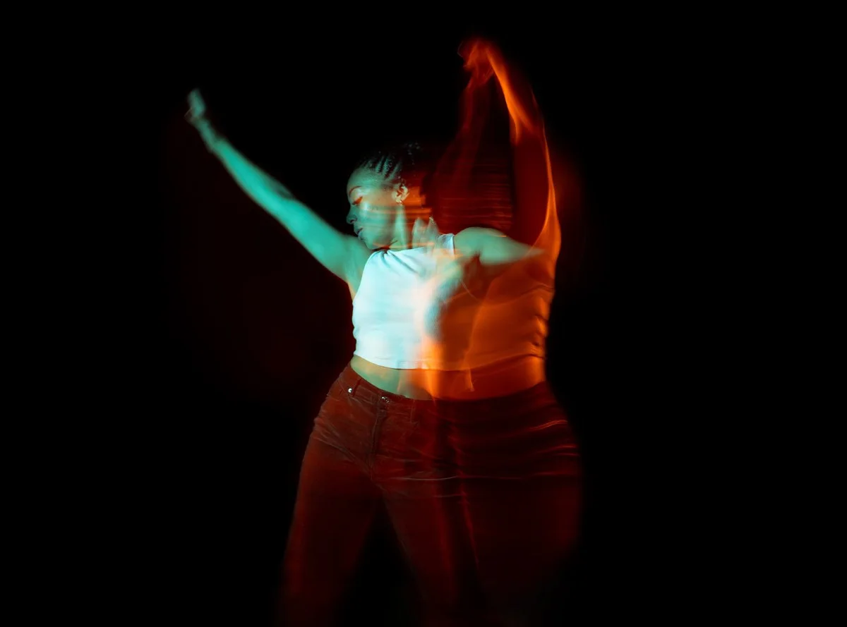 Mujer con top blanco sin mangas bailando sobre un fondo negro, el movimiento crea un efecto de doble exposición.
