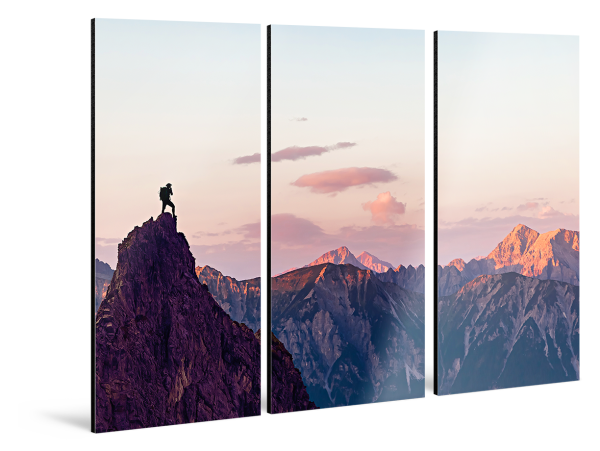 A man is standing on a rocky outcrop in front of a mountain panorama. The whole picture is divided into three parts hanging horizontally on the wall. 