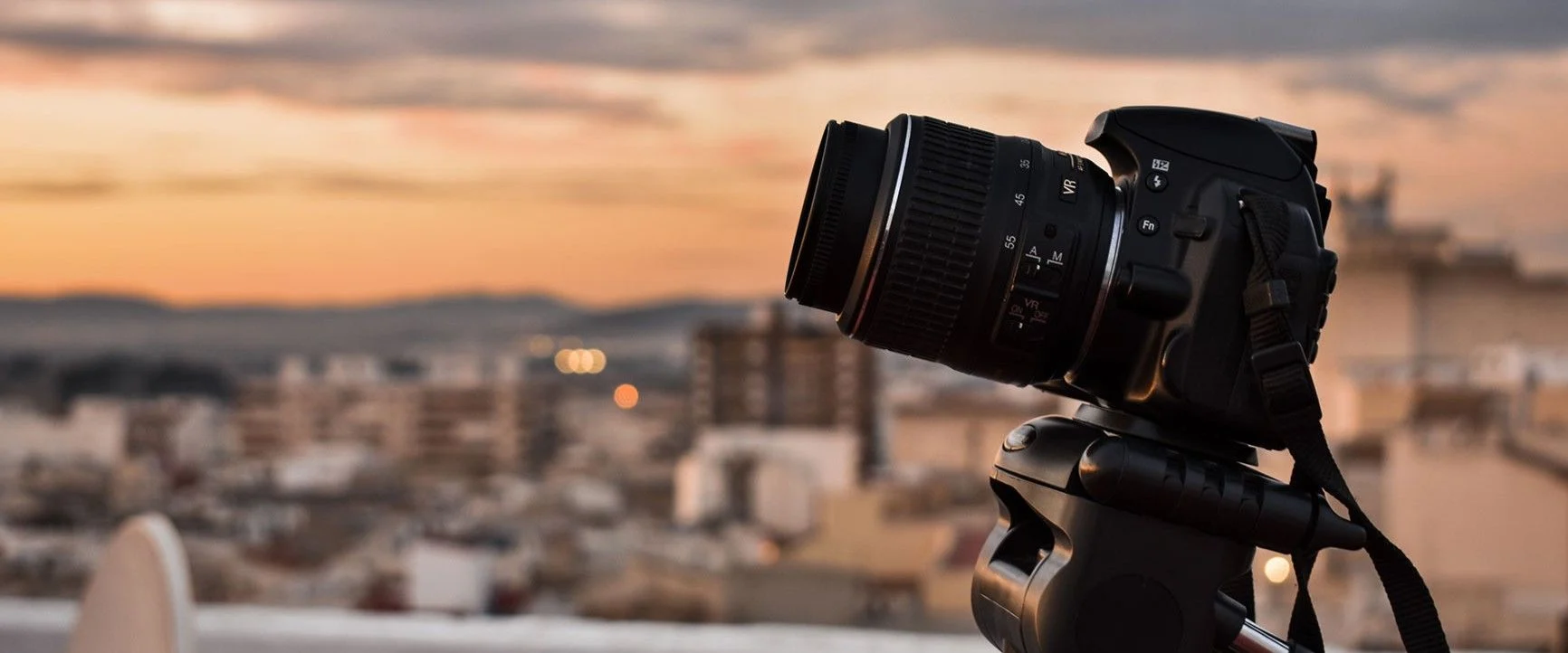A digital camera with a lens mounted on a tripod, set against a blurred cityscape at sunset.