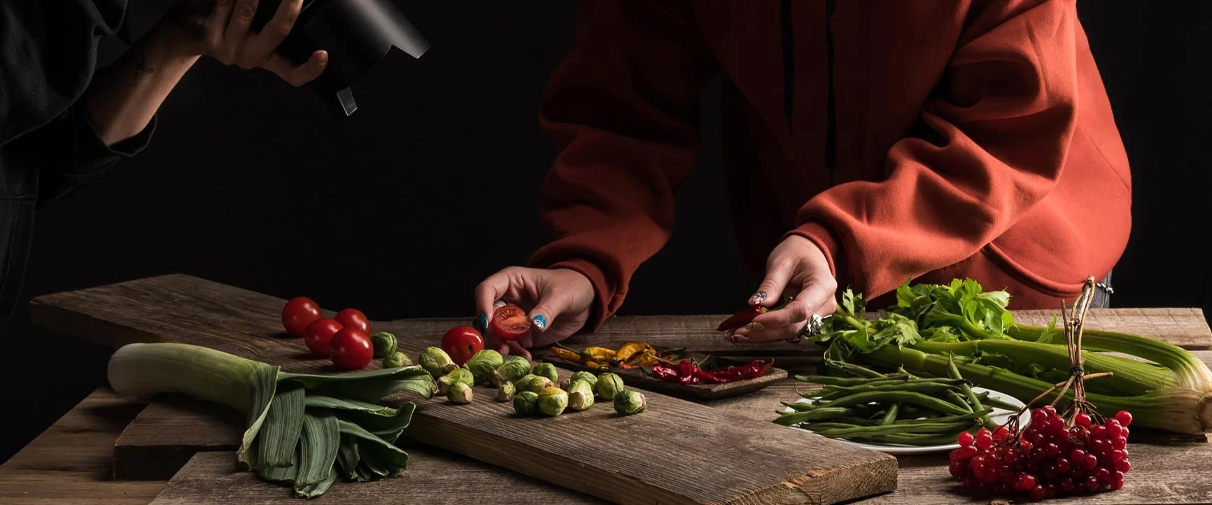 A person in a red sweater is arranging vegetables on a wooden board in front of another person holding a camera in a dark room.