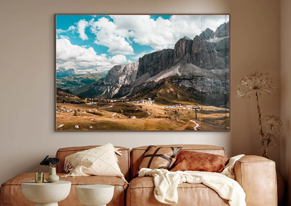 A large landscape painting with mountains and clouds hangs above a beige-brown leather sofa with decorative cushions and a blanket.