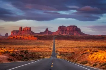 A straight road leads through red desert landscape to striking rock formations under a cloudy sky.