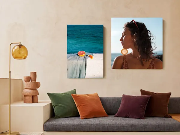 Interior de salón moderno y minimalist con dos impresiones artísticas verticales montadas en una pared beige de tono cálido sobre un sofá de tela gris. La impresión izquierda muestra un pomelo cortado apoyado sobre un paño azul claro con un fondo de océano turquesa, mientras que la impresión más grande a la derecha presenta un retrato de perfil de una mujer sosteniendo una rodaja de cítrico junto a su barbilla. A la izquierda del sofá, una lámpara de pie dorada se encuentra al lado de una escultura de terracota sobre un saliente de hormigón, und el sofá está decorado con cojines de terciopelo verde, naranja, morado y marrón.