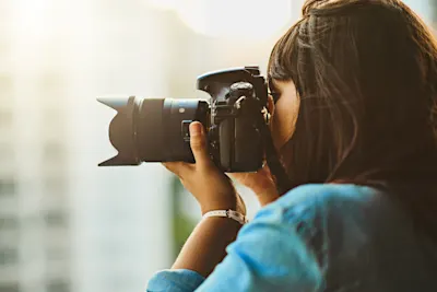 Frau mit langen dunklen Haaren hält eine DSLR-Kamera zum Fotografieren nah an ihr Gesicht.