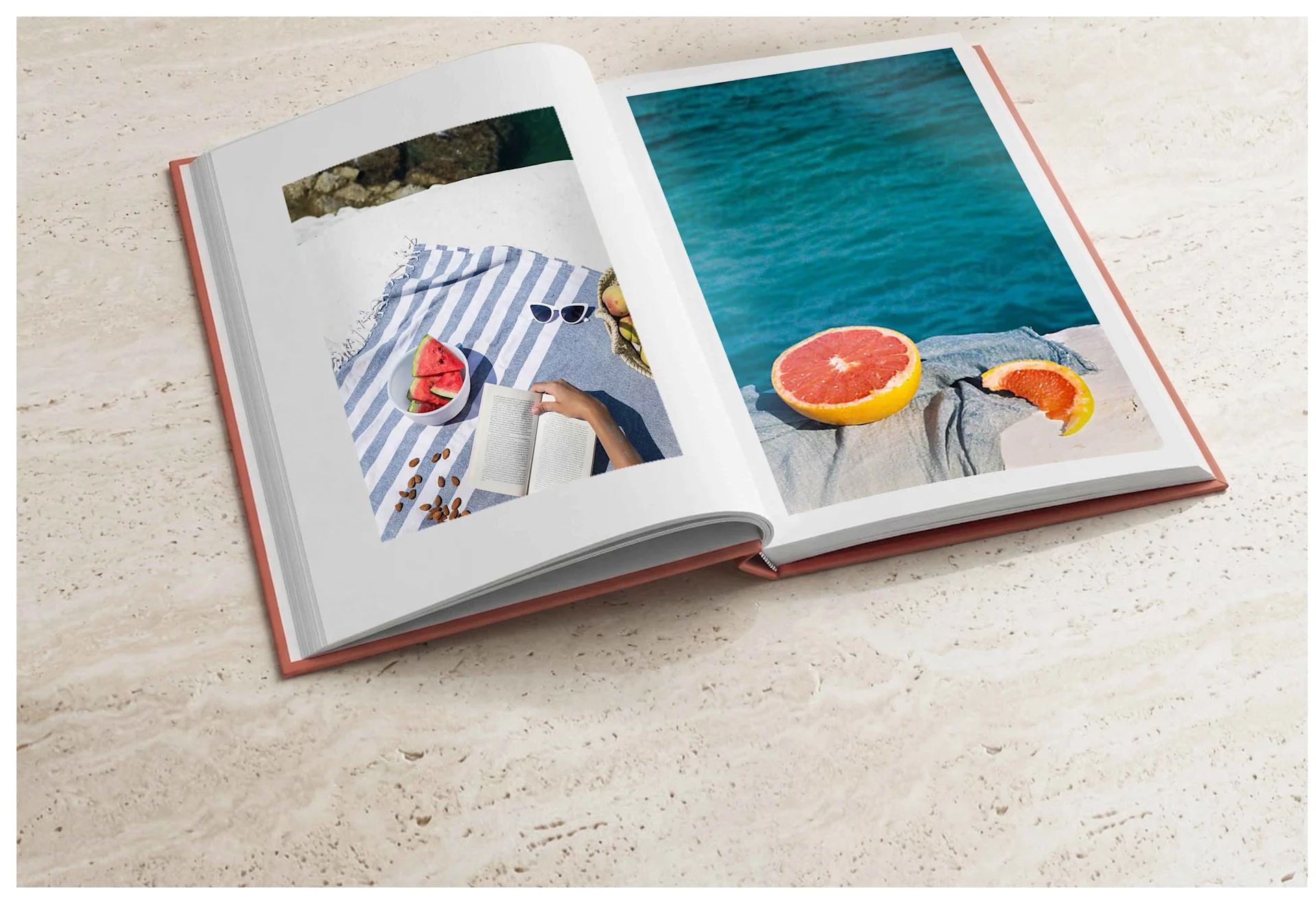 Un libro de arte abierto con una cubierta de color terracota descansa sobre una superficie de piedra crema texturizada. La página izquierda muestra una fotografía cenital de un pícnic sobre una manta a rayas azules und blancas con un cuenco de sandía, almendras y una persona leyendo. La página derecha presenta una fotografía a sangre de un pomelo cortado junto al océano turquesa. Una luz suave proyecta sombras sutiles que resaltan la calidad de impresión premium.