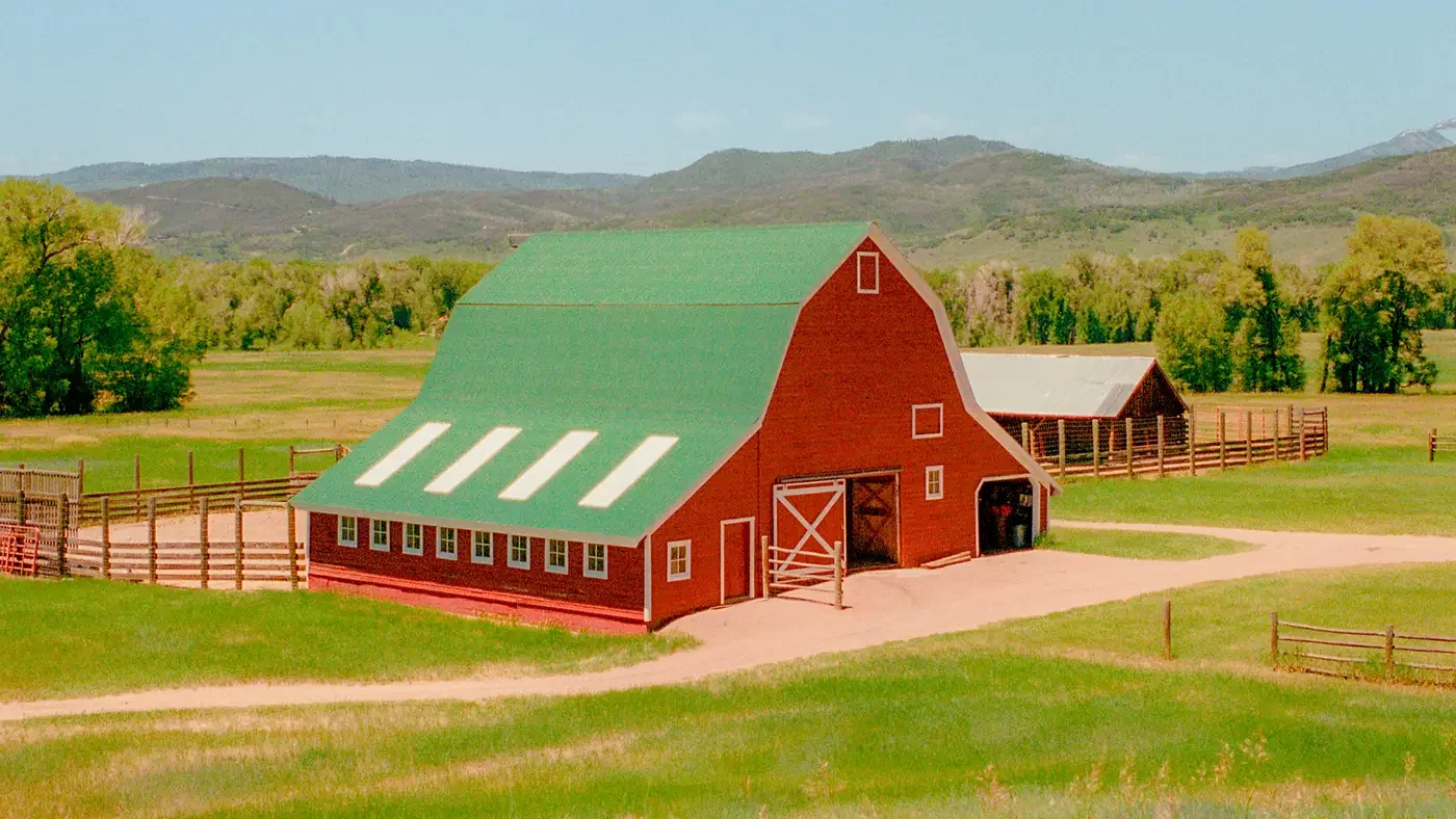 Rotes Bauernhaus mit grünem Satteldach auf einer Wiese vor bewaldeten Hügeln.