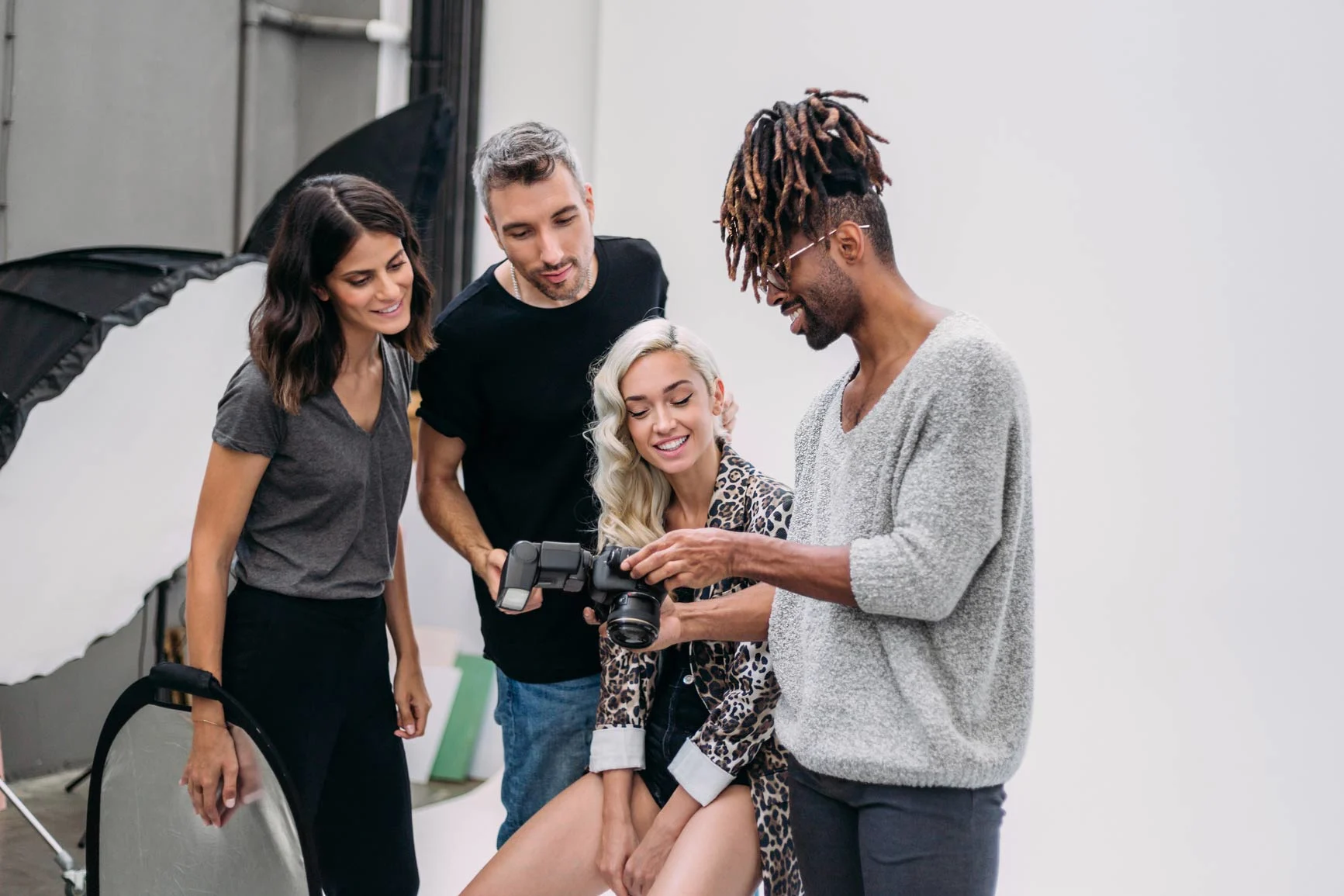 Four people are standing in a photo studio. One person is holding a camera and showing the display to the others. Studio lights are visible in the background.