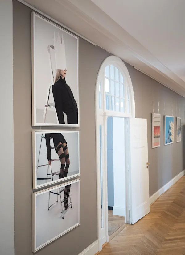 A law-firm hallway with framed fashion photos of a woman in black on a ladder, beside an arched white window and herringbone wood floor.