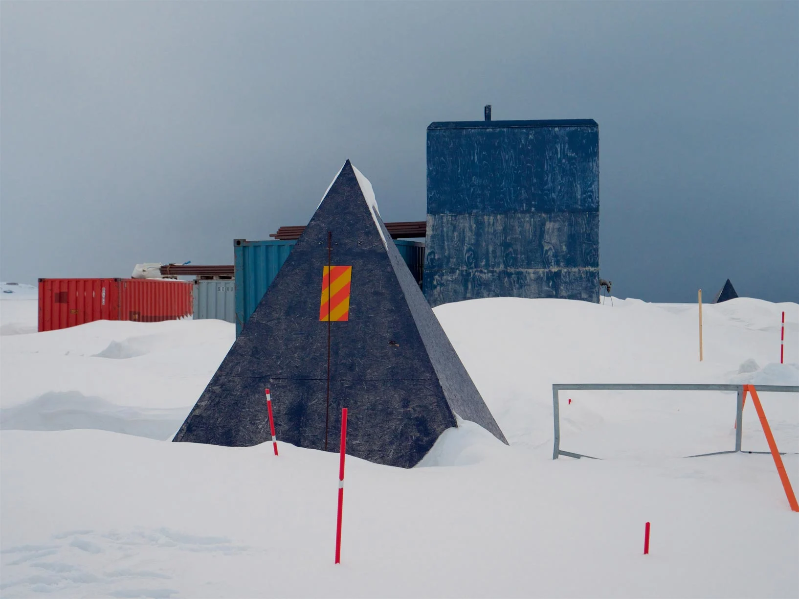 Estructura piramidal oscura, contenedores y edificios rectangulares se alzan en un paisaje nevado con postes indicadores.