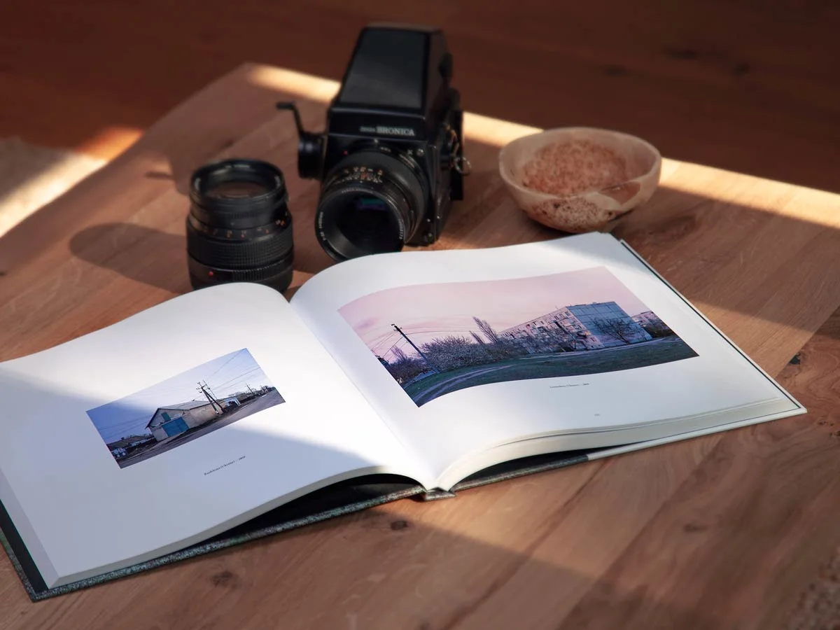 A photo book lies open on a wooden table and shows two landscape photographs. Next to the book are a camera, a camera lens and a bowl.