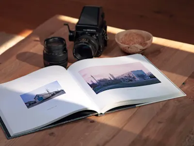 Open photo book on a wooden table, camera, two lenses and a bowl next to it. Light falls on the scene.
