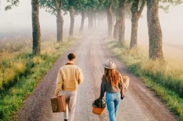 Two people with luggage walk along a tree-lined gravel road through a misty landscape.