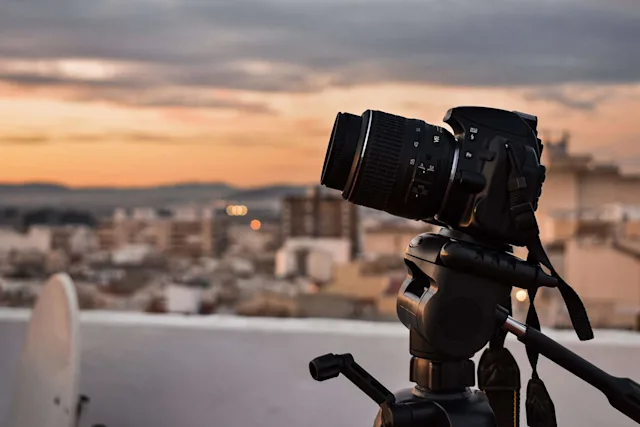 A digital camera with a lens mounted on a tripod, set against a blurred cityscape at sunset.