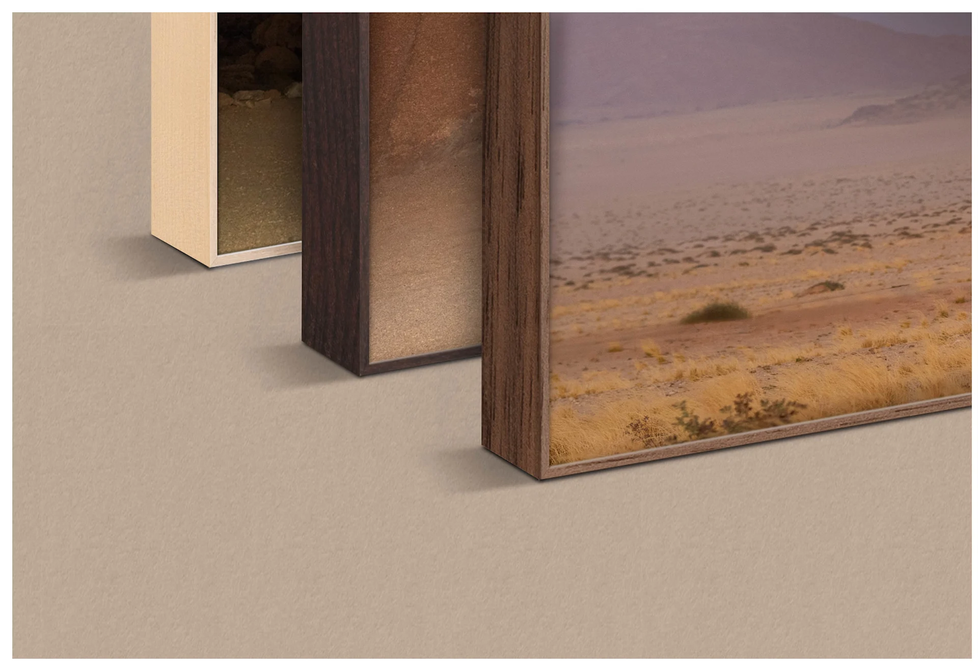 Un gros plan montre trois cadres ArtBox côte à côte dans différentes finitions de bois. De gauche à droite, on aperçoit une finition en érable clair, une finition en noyer foncé et un cadre en aspect chêne. Chaque cadre contient une partie du même motif de désert avec une caravane Airstream argentée sous un ciel violet. Les cadres sont posés sur une surface neutre de couleur sable.