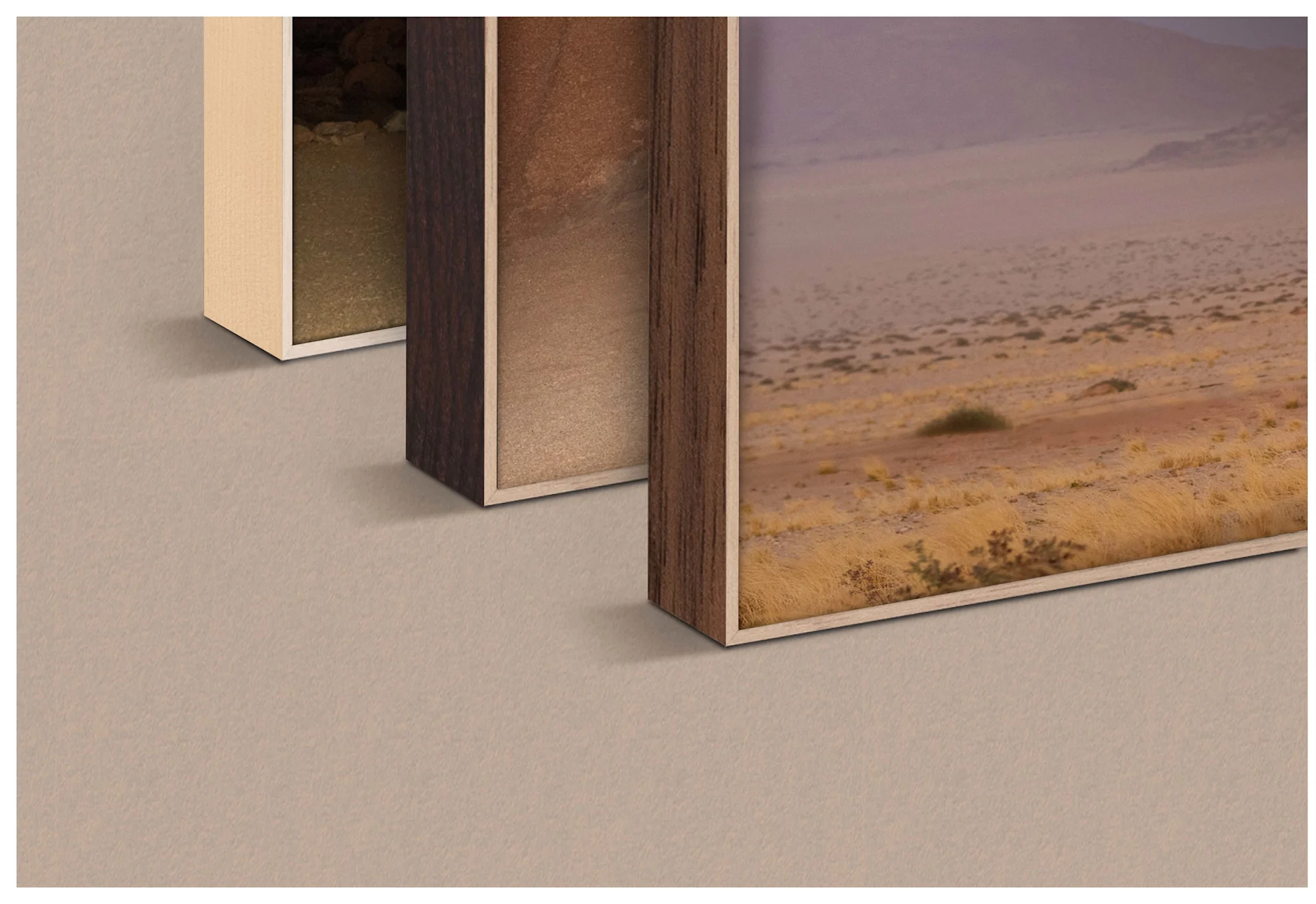 A close-up shot shows three ArtBox frames side by side in different wood finishes. From left to right, a light maple finish, a dark walnut finish, and an oak-look frame are visible. Each frame contains a section of the same desert motif featuring a silver Airstream trailer under a purple sky. The frames stand on a neutral, sand-colored surface.